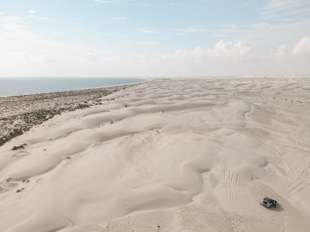 Aerial photo of white car parked in expanse of white sand dunes which run down to the beach and a turquoise sea at the Sugar Dunes, Oman