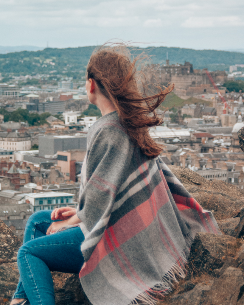 Woman in tartan scarf sat on Arthur's Seat with view of Edinburgh Castle