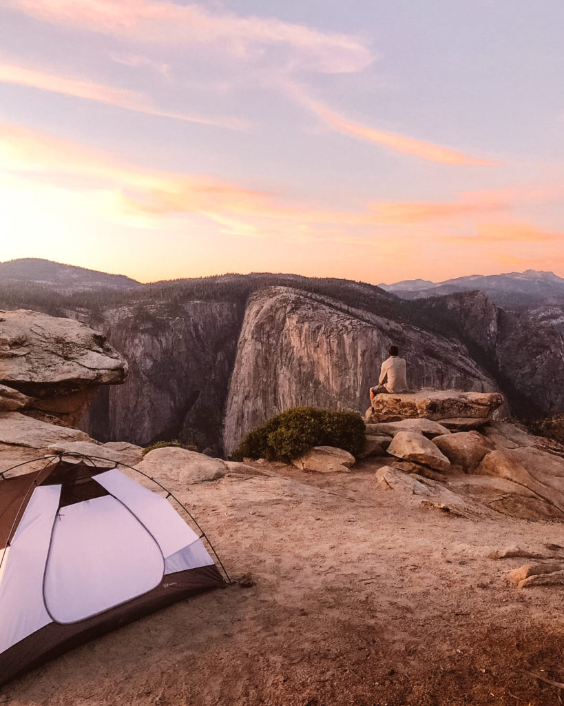 Man sat on rock overlooking El Capitan at sunset with tent set up for wild camping in Yosemite