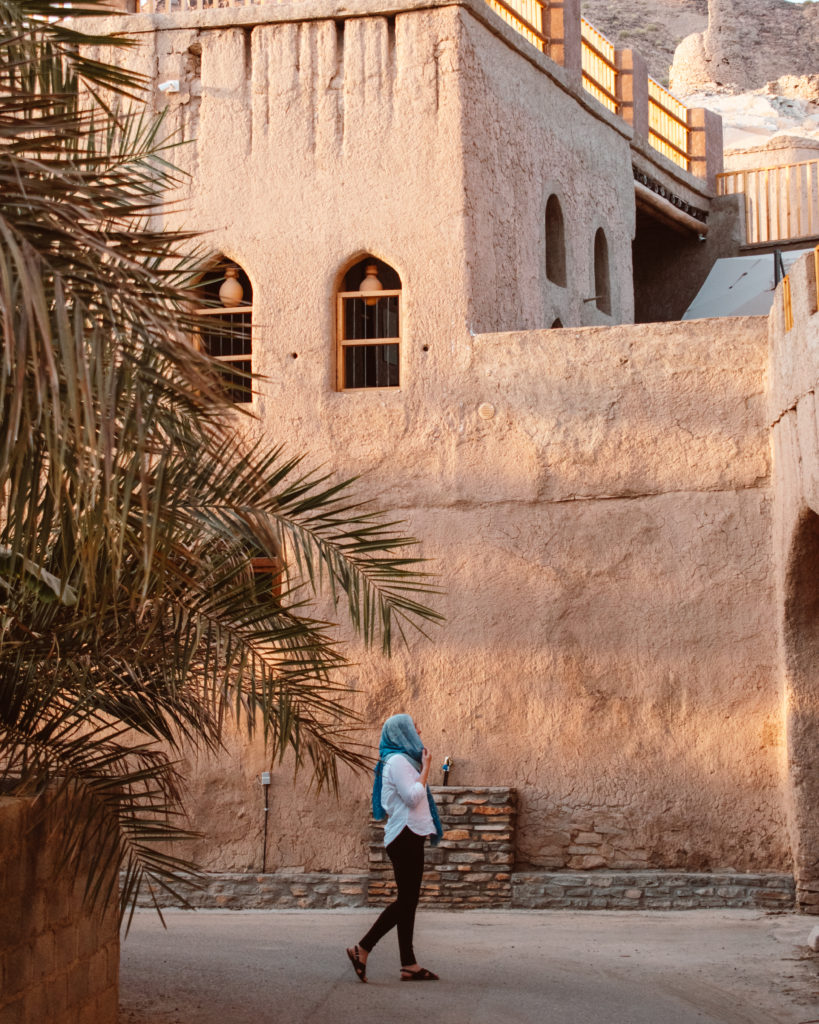 Woman in blue headscarf walking in front of old building in Birkat Al Mouz