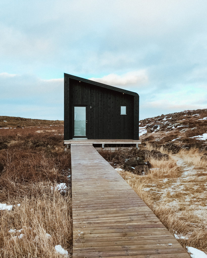 Wooden path on snowy grass leading to an asymmetric dark wooden cabin 