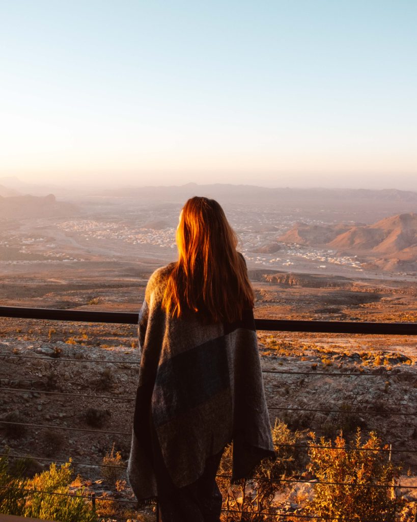 Woman wrapped in plaid blanket looking at sunrise over Al Hamra, Oman