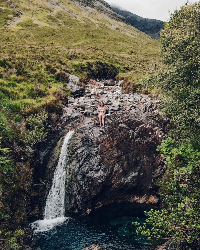 Woman in pale swimsuit sat at the top of a waterfall, in front of rugged green mountains