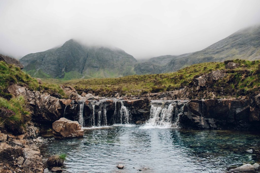 Fairy Pools: Small waterfalls flowing into a blue pool of water in front of mist covered mountains