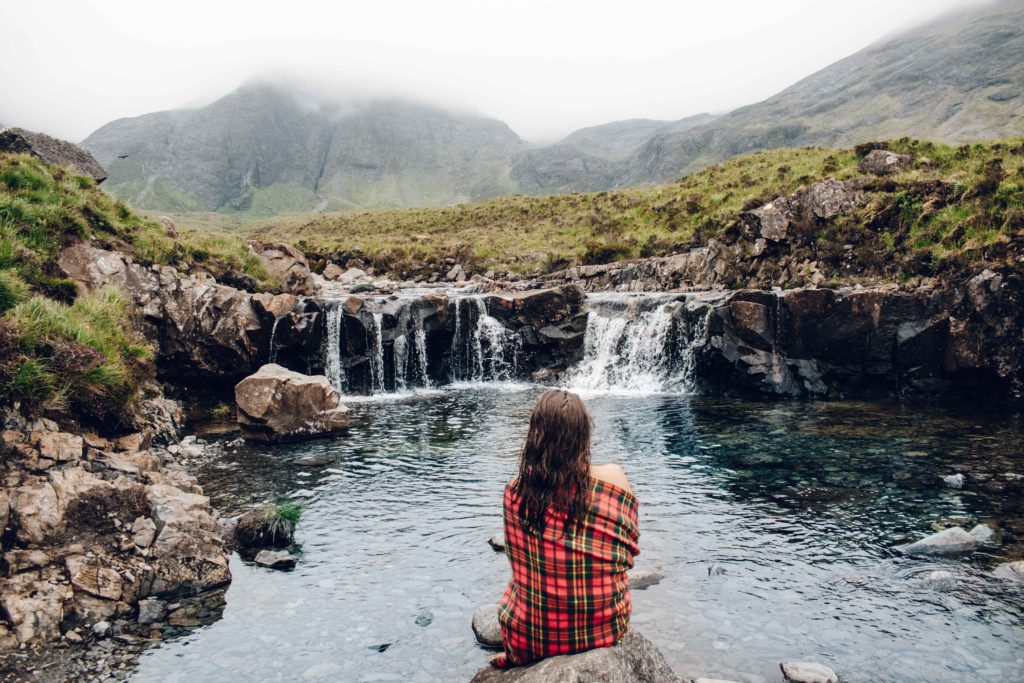 Woman wrapped in tartan blanket looking at small waterfalls and mountains beyond in Scotland 
