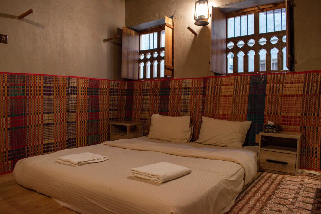 Mattress covered with cream bedding on a floor in a room with red traditional Omani fabric on the walls, and decorative wooden blinds  on the windows