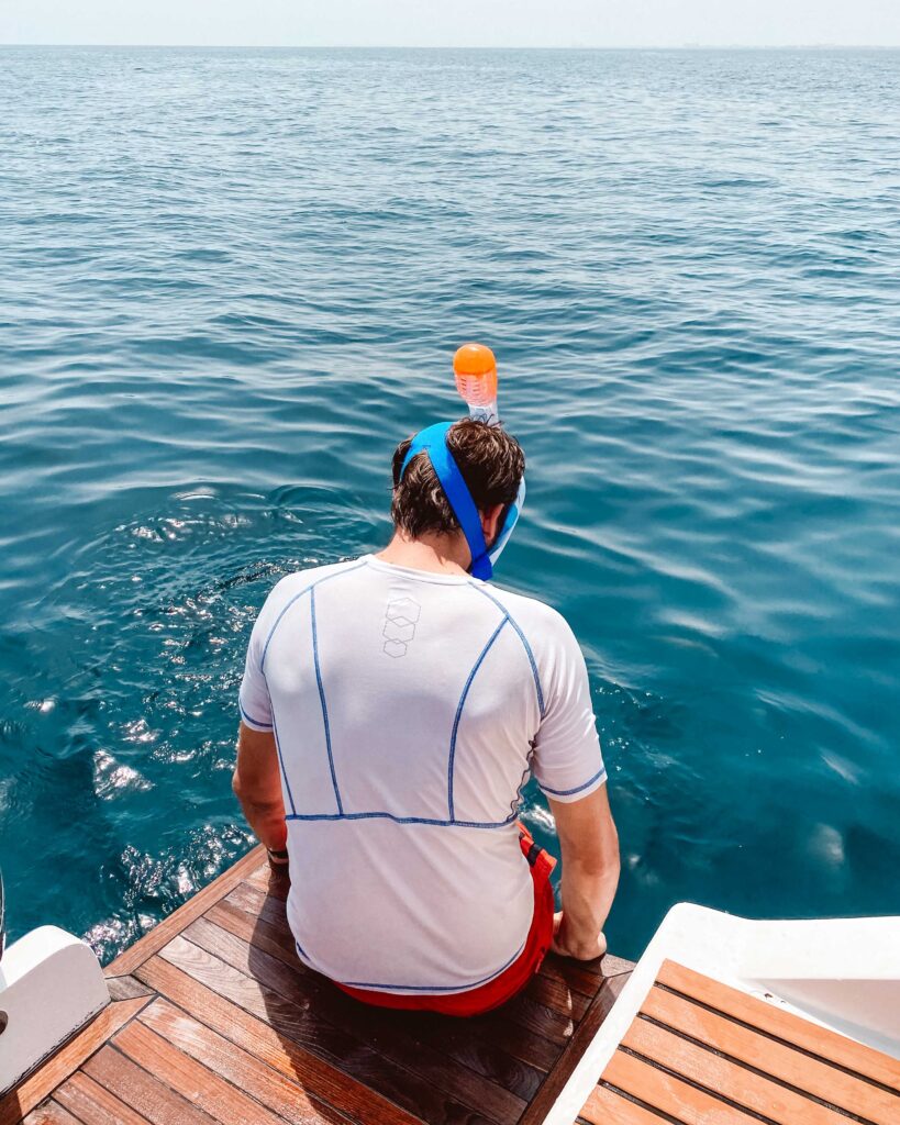 Man in white t-shirt and blue snorkel mask sat on edge of a boat in the Daymaniyat Islands