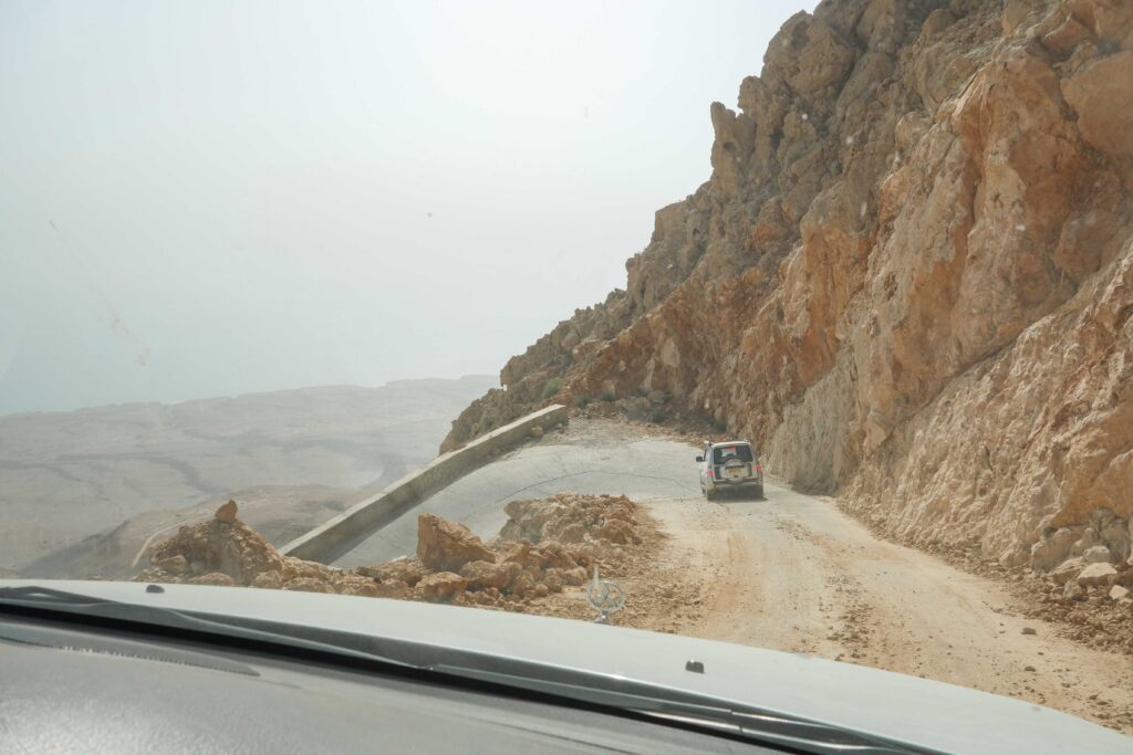 Car on narrow road underneath rocky mountains descending the Salma Plateau, Oman