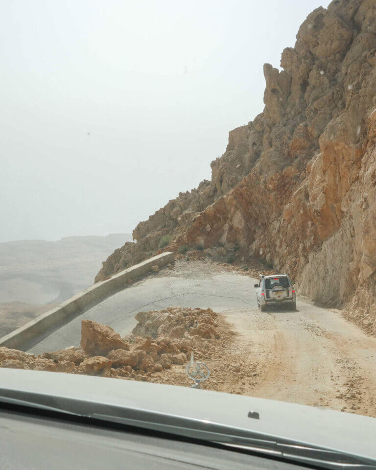 Car on narrow road underneath rocky mountains descending the Salma Plateau, Oman