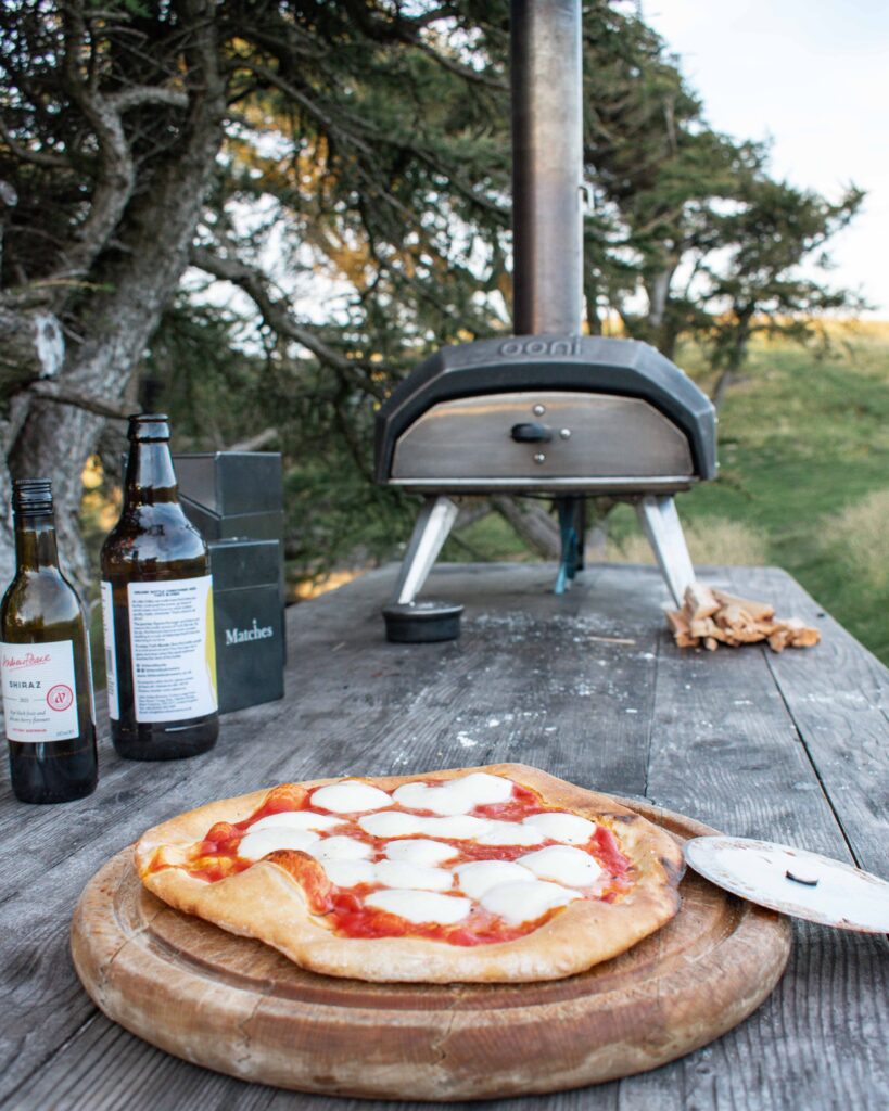 Freshly made pizza on wooden board in front of Ooni pizza oven on outdoor table, at Hinterlandes Hidden Hut
