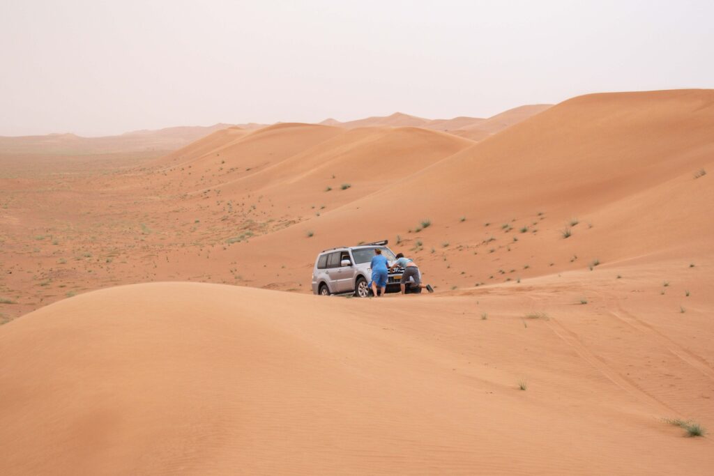 Man and woman pushing a silver car on a sand dune in Wahiba Sands, Oman