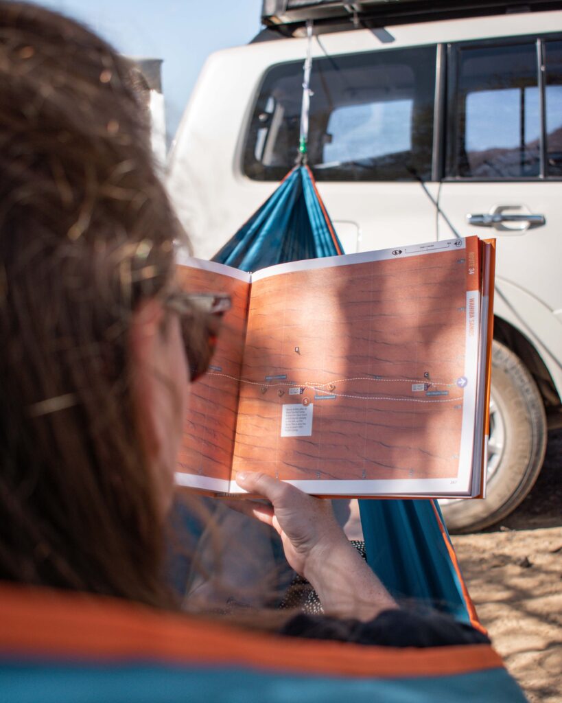 Woman in blue hammock string from a car reading a route description in the Oman Off Road guidebook
