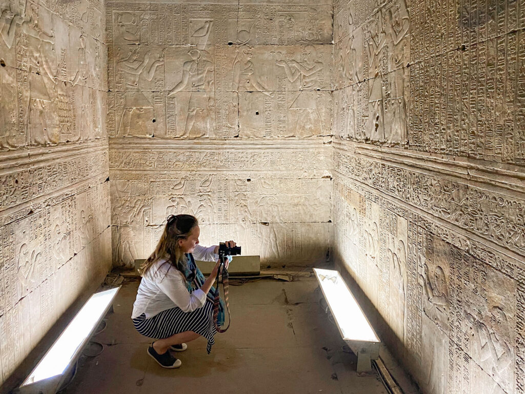 Woman in white shirt knelt down photographing intricately carved walls of the incense room at Edfu Temple, Egypt