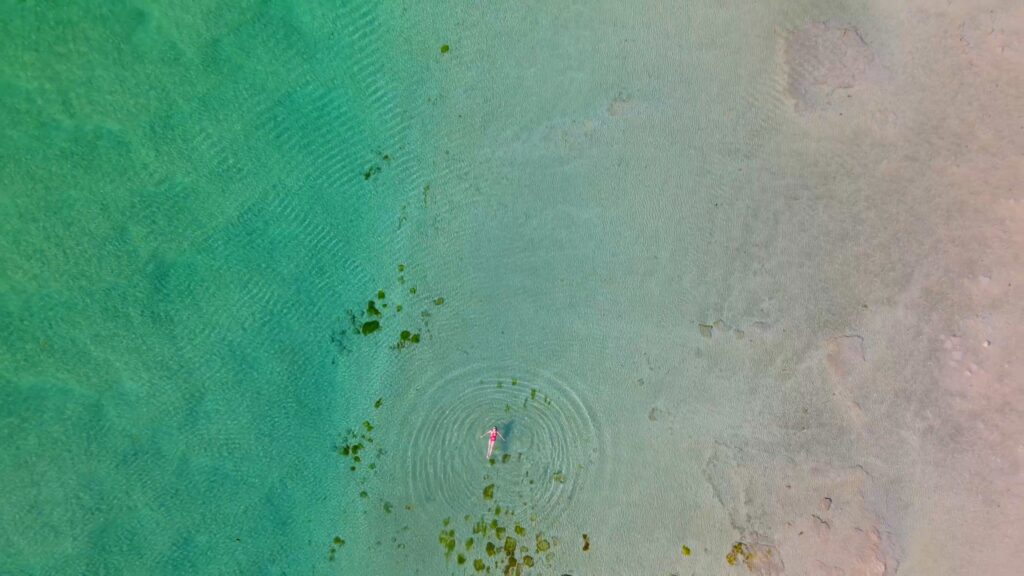 Woman in a red swimming costume floats on her back in crystal clear, turquoise water in a drone photo above Luskentyre Beach in the Outer Hebrides. 