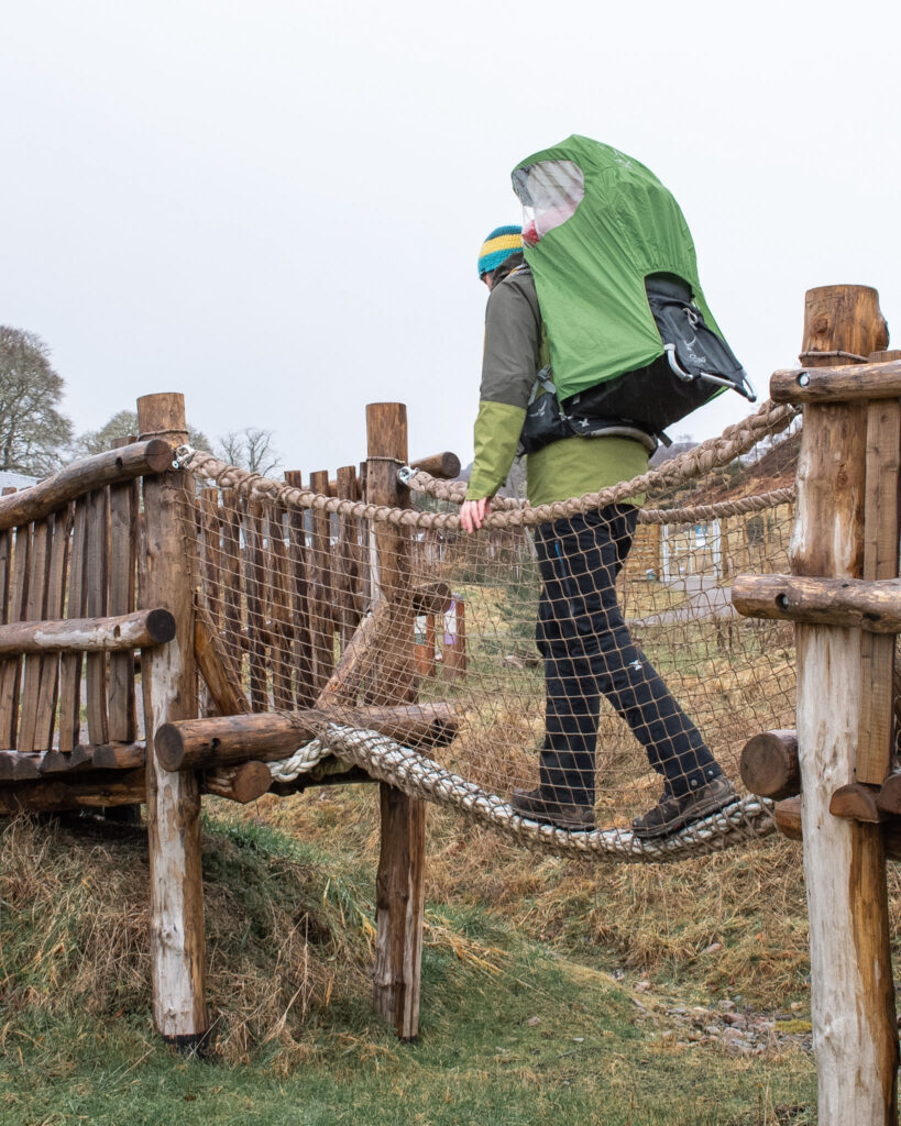 A man in black waterproof trousers and a green waterproof walks across a rope bridge as part of a wooden climbing frame, whilst carrying an Osprey Poco Plus baby carrier with green rain cover at Dundreggan Rewilding Centre. 