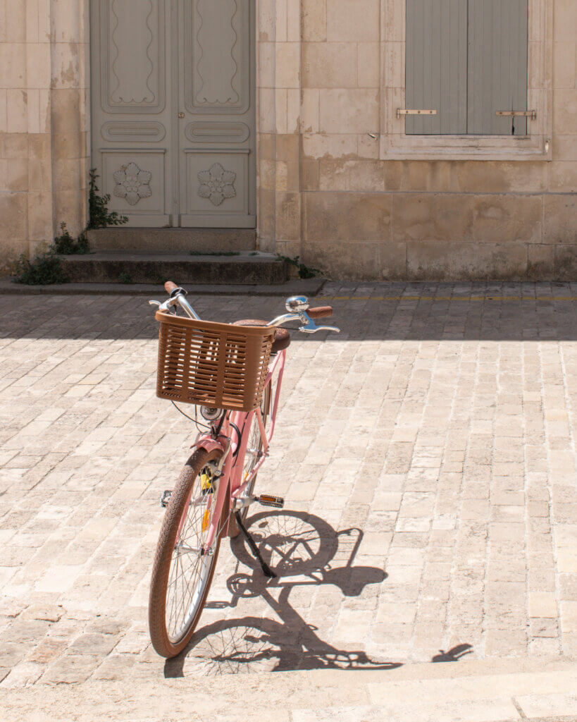 Pink bicycle with basket on the front, is parked in the sunshine in front of a stone building with pale grey shutters in Ars-en-Re.