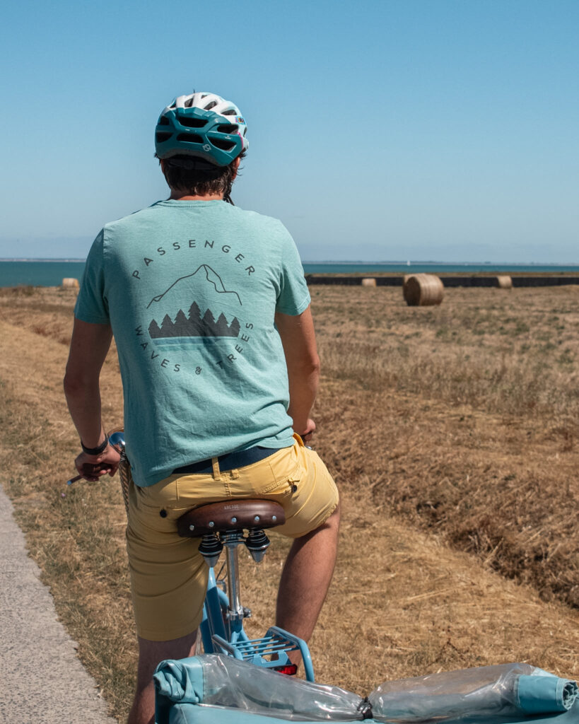 A man in a green Passenger Clothing t-shirt and yellow shorts sits on a stationary bike, looking out over a field of straw bales on the Ile de Re.