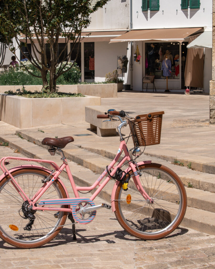 A pink bike with basket is parked at the bottom of steps in the centre of a village on the Isle de Re.
