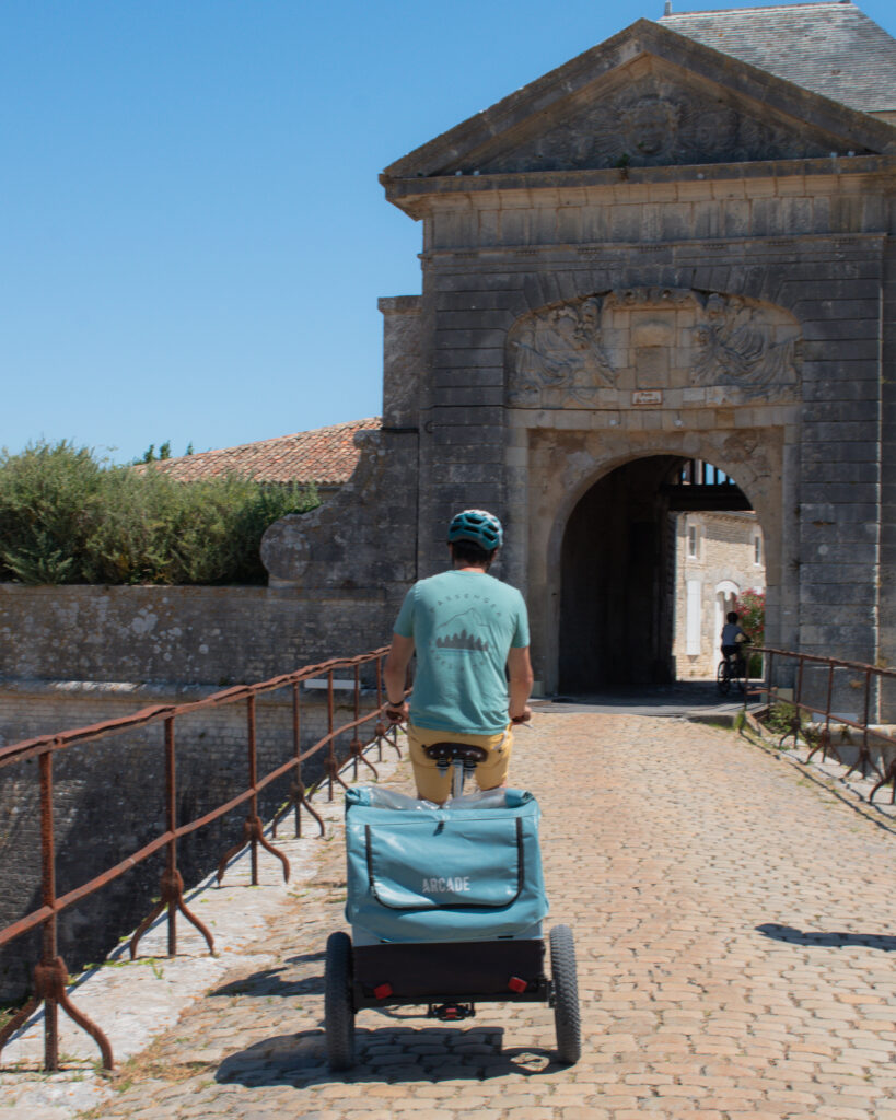 A man in a green t-shirt cycles under the historic stone archways, to enter Sain-Martin-de-Re, pulling a children's bike trailer.