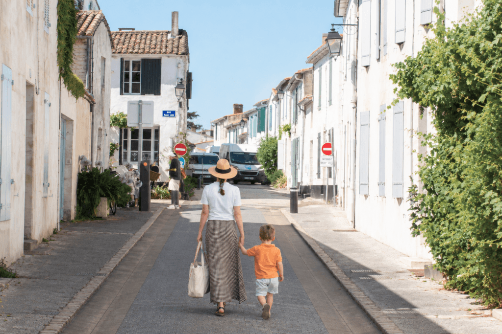 Woman in summer clothing holds the hand of a small child, walking away from the camera in the sunny streets of La Flotte.