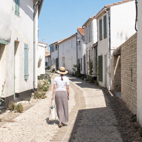 A woman in a white tshirt and summer skirt, walks down a sunny street in Saint Martin de Re.
